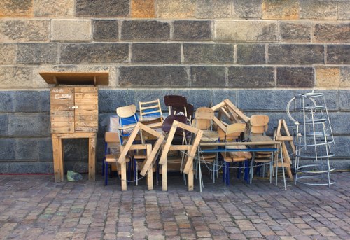 A collection of various wooden chairs and stools, stacked and arranged haphazardly, positioned against a cinder block wall outdoors on a paved brick surface. The chairs include different styles, some with metal legs and others with wooden legs, with finishes ranging from natural wood to painted surfaces in shades of white, blue, and dark brown. In the background, there is a tall wooden cabinet or storage unit with a slanted top, and to its right, a metal or wire shelving unit with multiple tiers. The scene appears to be in an outdoor urban or industrial area, possibly a site for rubbish removal or waste clearance, aligned with the services of Commercial Waste Removal Sutton. The overall setting is well-lit, and the objects are scattered, suggesting they are ready for collection or disposal.