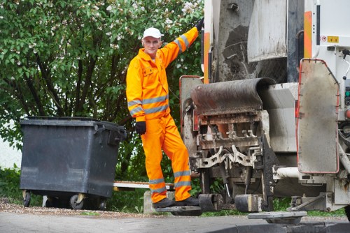A waste collection worker dressed in high-visibility orange and reflective striped safety clothing stands on a pavement next to a large black rubbish bin. The worker is positioned to the left of a commercial rubbish collection vehicle, which is partially visible with its mechanical bin lift and roller mechanism on the right side of the image. The background features leafy green trees, indicating an outdoor urban or suburban environment, possibly within or near Sutton. The worker is smiling and appears to be in the process of managing waste removal, aligning with the services offered by Commercial Waste Removal Sutton. The scene is well-lit, suggesting daytime, and the focus is on the interaction between the worker and the waste collection process, emphasizing the professional and efficient rubbish removal activity typical for local commercial waste services.