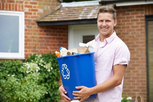 A young man with short, light brown hair and a friendly smile is standing outdoors in front of a red-brick building with a small window and garden plants. He is wearing a light pink short-sleeved shirt and is holding a large blue plastic recycling bin with a white recycling symbol on its side, filled with various waste items such as paper, cardboard, and plastic bottles. The background includes green shrubbery and flowering plants, indicating a residential area. The scene suggests a rubbish collection or waste disposal activity typical of homeowners or local waste management services in Sutton, highlighting a clean and organized environment related to rubbish removal services. The natural daylight enhances the clear, professional atmosphere of the image, aligning with the context of waste management and rubbish removal in the local area.