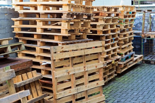 A large stack of wooden pallets is arranged neatly on a paved outdoor area, with several pallets leaning or partially stacked in front. The pallets are light-colored, made of treated timber with visible grain, and show varying degrees of weathering, some with darker marks and signs of use. The background includes a chain-link fence, some metal storage containers, and elements of an industrial or commercial yard, suggesting an environment related to waste management or logistics services. The scene is well-lit, indicating daylight, and the overall setting appears organized, with the pallets prepared for collection or recycling as part of a rubbish removal process. This image supports the context of commercial waste removal services provided by Commercial Waste Removal Sutton, situated in the Sutton area, focusing on the handling and disposal of wooden packaging materials.