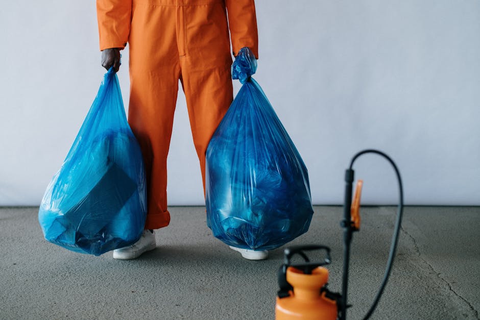A person wearing orange protective overalls and white footwear is standing on a concrete floor, holding two large blue plastic rubbish bags filled with waste, with the bags appearing opaque and tightl